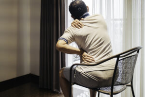 Um homem sentado, aparentemente em desconforto, segurando sua lombar e pescoço, representando sintomas comuns de lesão muscular crônica