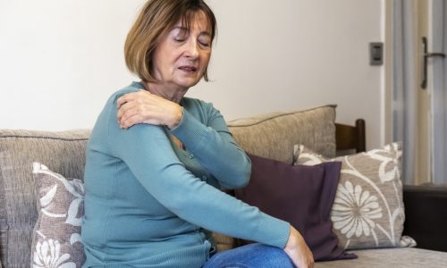 Mulher idosa sentada no sofá e tocando o ombro devido a artrose no ombro
