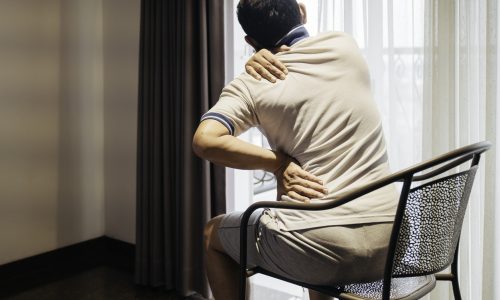 Um homem sentado, aparentemente em desconforto, segurando sua lombar e pescoço, representando sintomas comuns de lesão muscular crônica